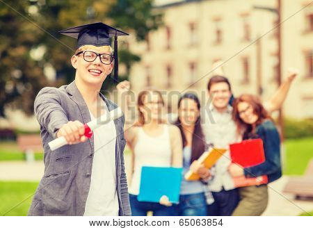 education, campus and teenage concept - smiling teenage boy in corner-cap and eyeglasses with diploma and classmates on the back