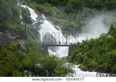 Tourists in water shower on the bridge of Briksdalsbreen hike waterfall. Norway popular touristic hike to Briksdal glacier.