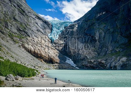 Tourists at Briksdalsbreen glacier viewpoint. Rocky mountains, melted ice lake and gravel beach. Popular travel destination of Norway.