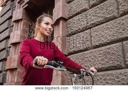 Joyful young woman dressed in sweater walking with her bicycle near wall on the street and smiling. Look aside.