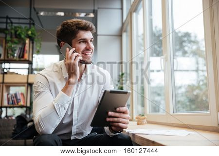 Smiling businessman dressed in white shirt sitting in cafe and using tablet computer while talking by his phone. Look aside.