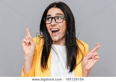 Funny woman wearing eyeglasses and dressed in yellow jacket crossed fingers over grey background. Look aside.