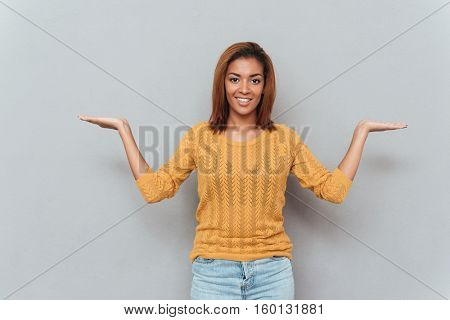 Smiling african woman in yellow sweater and jeans choosing between copyspace as well as looking at camera. Isolated gray background