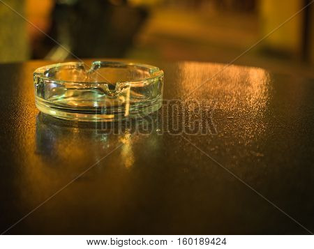 Picture of the glassy ashtray close up. Ashtray laying on the blurred surface of the table. Blurred background of the ashtray on the table. Blurred surface of the table reflects yellow sunlight.