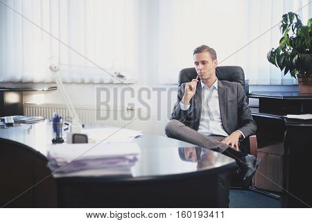 Male adult man in a suit sitting on a chair behind the glossy reflective desk in a office, looking down with pen next to his cheek, thinking thoughtful