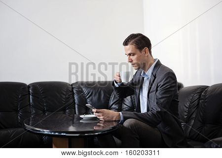 Male adult businessman sitting behind smal oval dark shiny table on a black leather sofa. Man is holding coffee cup in one hand and cellphone in other hand. Looking down on the phone.