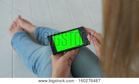 Woman sitting on the white wooden floor and using horizontal smartphone with green screen. Close up shot of woman's hands with mobile Close up shot of woman's hands with mobile