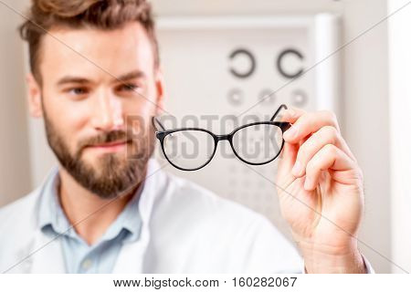 Handsome ophthalmologist looking at the glasses in front of the eye chart in the cabinet. Image with small deph of field focused on hands and glasses