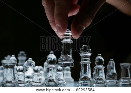 Close-up view on the transparent glass chess board with male hand holding one of the chess pieces. Black background.