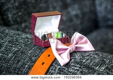 The groom's sophisticated accessories including cuff links in the box leather brown belt and pink bow tie placed on a grey sofa