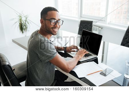 Image of cheerful african man dressed in gey t-shirt and wearing eyeglasses using laptop and sitting at the table. Looking at the camera.
