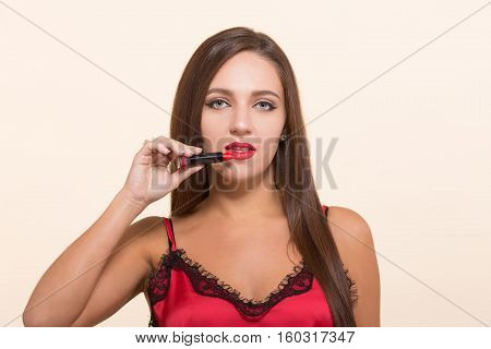 Serious young model woman holding lipstick near her lips and looking at camera while posing for photographer isolated on white background.