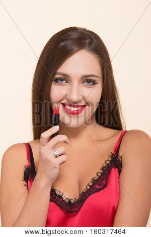 Young model woman happy smiling for camera while holding red lipstick near her face and lips. Red lipstick for doing makeup isolated on white.