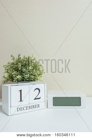 White wooden calendar with black 12 december word with clock and plant on white wood desk and cream wallpaper textured background selective focus at the calendar