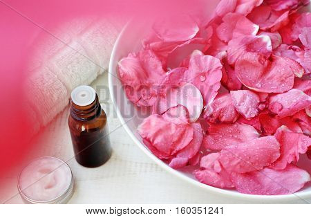 Aromatherapy body treatment, warm herbal bath soak. Bowl of Rose petals, essential oil, complimentary emollient sample. Luxury natural skincare line.  Framed natural pink flower blur.