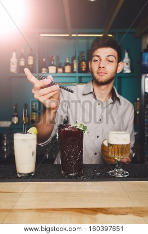 Young handsome Barman offers exotic sweet cocktails in restaurant. Professional bartender at work in night club bar made refreshing drinks. Selective focus on glasses
