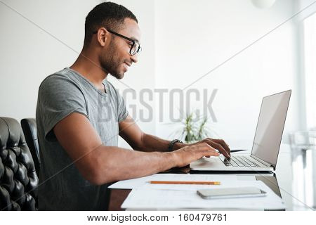 Happy african young man dressed in gey t-shirt and wearing eyeglasses using laptop and sitting at the table.
