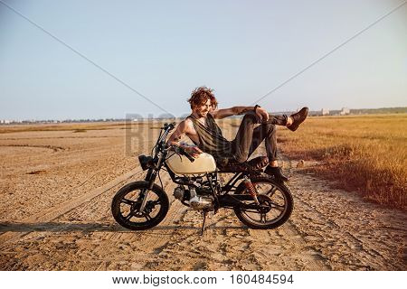 Young brutal man laying on his motorcycle in the desert and posing