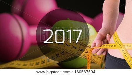 Mid section of woman measuring her waist against 2017 with dumbbells in background