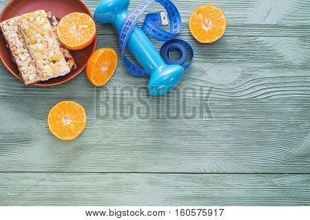 The concept of healthy lifestyle top view. Juicy citrus cereal bars dumbbell and measuring tape on green wooden table flat lay. Cereals and fruits - diet and breakfast
