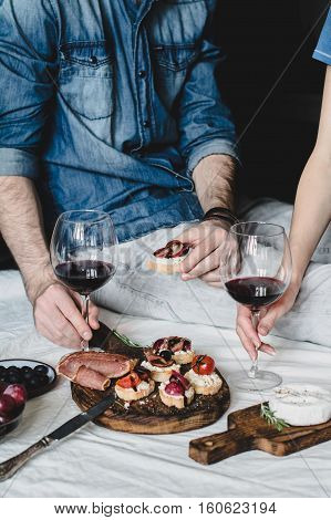 Couple eating dinner with red wine and antipasto snacks set: bruschettas, crostini, prosciutto, camambert cheese, grapes and black olives. Selective focus, desaturated effect.