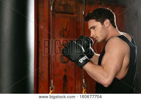 Picture of young strong boxer training in a gym with punchbag. Looking at punchbag.