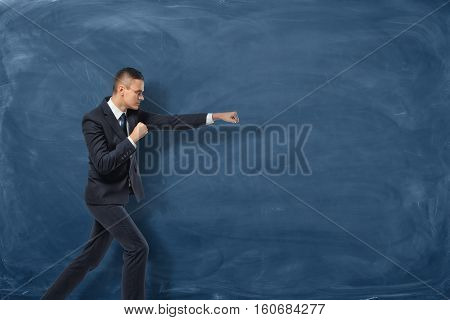 A businessman in a black suit standing as if he is going to punch something invisible near the blue chalkboard. Aggressive marketing strategies. Business development. Successful leader.