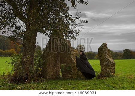 Donna o una strega nel mantello nero camminare intorno ad un gruppo di menhir e un albero solitario