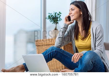 Involved in the conversation over the phone. Delighted happy cheerful woman sitting on the floor in front of the laptop while speaking over the phone and expressing happiness