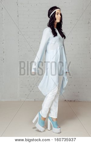 Model in studio posing to the photograther staying near grey bricks wall. She has beautiful white costume
