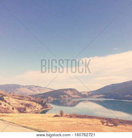 Scenic lake and mountains landscape view with calm blue water and reflections of mountains on lake.Bright clear blue sky and white cloud. Field of dry grass and shrubs and bushes In foreground.