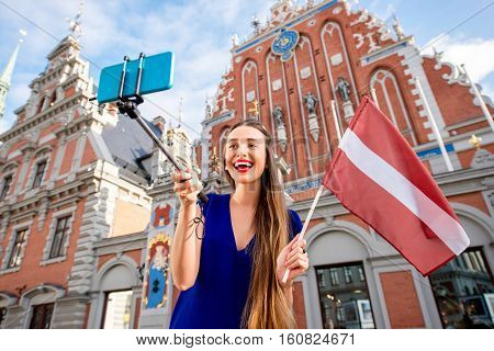 Young female tourist making selfie portrait with latvian flag on the old town square in Riga. Woman having great vacations in Latvia