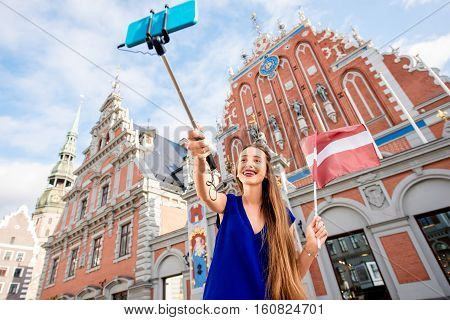Young female tourist making selfie portrait with latvian flag on the old town square in Riga. Woman having great vacations in Latvia