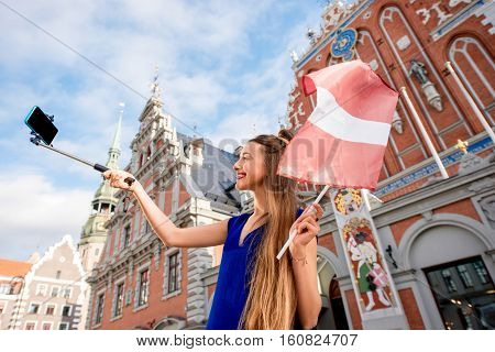 Young female tourist making selfie portrait with latvian flag on the old town square in Riga. Woman having great vacations in Latvia