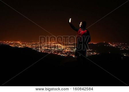 Young man with phone on top of the hill observing the night city view.