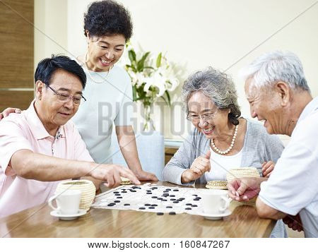 two senior asian men playing Weiqi (or game of go) at home with their wives watching.