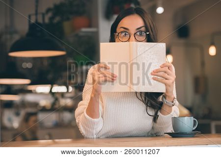 Shocked Woman In Eyeglasses Covering Face By Book At Table With Coffee Cup In Cafe