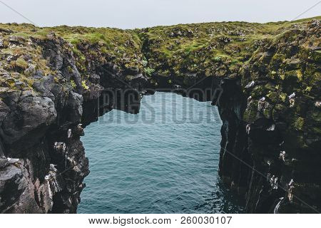 Mossy Rocky Cliff In Shape Of Arch With Blue Ocean On Background In Arnarstapi, Iceland