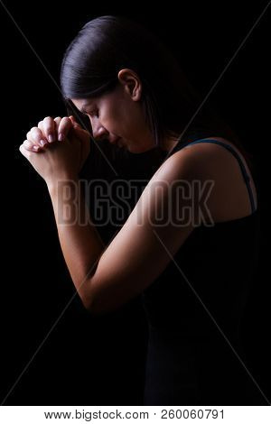 Faithful woman praying, hands folded in worship to god with head down and eyes closed in religious fervor, on a black background. Concept for religion, faith, prayer and spirituality.