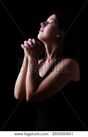Faithful woman praying, hands folded in worship to god with head up and closed eyes in religious fervor, on a black background. Concept for religion, faith, prayer and spirituality.