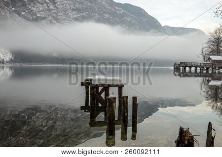 Decaying Wooden Pier In Winter Lake Bohinj With Mountains In Background And Myst Above The Water.