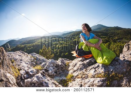 A Woman Is Sitting In A Sleeping Bag At Sunset. Camping On Top Of A Mountain. A Smiling Girl Rests A