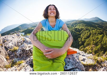 A Woman Is Sitting In A Sleeping Bag At Sunset. Camping On Top Of A Mountain. A Smiling Girl Rests A