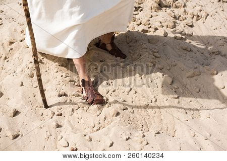 Cropped Image Of Jesus In Robe And Sandals Walking On Sand With Wooden Staff In Desert