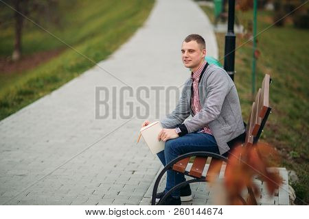 A Cute Student Sits On The Bench And Writes Down His Thoughts In His Notebool Using A Pensil. Study 