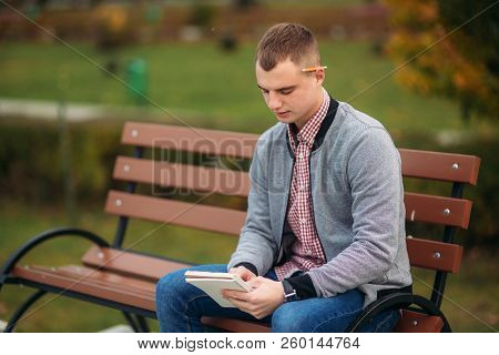 A Cute Student Sits On The Bench And Writes Down His Thoughts In His Notebool Using A Pensil. Study 