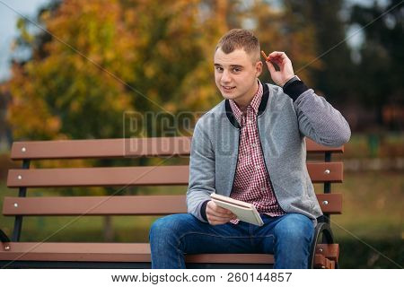 A Cute Student Sits On The Bench And Writes Down His Thoughts In His Notebool Using A Pensil. Study 