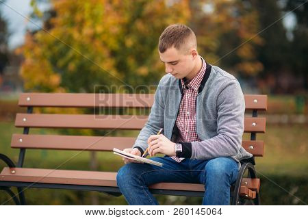 A Cute Student Sits On The Bench And Writes Down His Thoughts In His Notebool Using A Pensil. Study 
