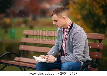 A Cute Student Sits On The Bench And Writes Down His Thoughts In His Notebool Using A Pensil. Study 