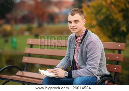 A Cute Student Sits On The Bench And Writes Down His Thoughts In His Notebool Using A Pensil. Study 